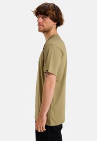 Camiseta de algodón verde oliva de manga corta, con un sutil logo en el pecho y una etiqueta en la manga. Diseño de corte recto.