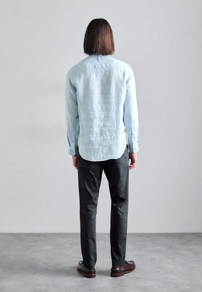 Camicia in lino azzurro chiaro, maniche lunghe, orlo curvo, abbinata a pantaloni scuri a sigaretta e scarpe in pelle marrone. Sfondo neutro.
