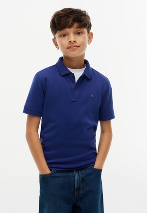 Garçon aux cheveux courts et foncés, portant un polo bleu et un jean, les mains dans les poches, debout devant un fond blanc.