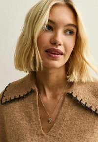 Maglione di lana marrone chiaro con scollo a V e colletto ricamato nero. Una collana d'argento presenta un ciondolo a forma di cuore. Texture morbida.