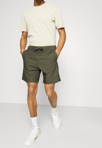 Homme portant un t-shirt à manches courtes crème, un short à cordon olive, des chaussettes blanches et des baskets blanches, marchant avec les mains dans les poches.