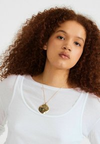 Jonge vrouw met krullend bruin haar, gekleed in een doorschijnend wit shirt en een grote, onregelmatige gouden hanger tegen een effen achtergrond.