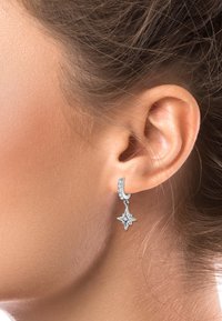 Silberne Creolen mit einem sternförmigen Anhänger, der mit klaren Strasssteinen verziert ist und eine polierte Oberfläche hat, auf einem Ohr präsentiert.