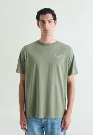 Hombre con cabello corto, rizado y oscuro, vistiendo una camiseta verde oliva con un pequeño logo claro en el pecho y vaqueros azul claro, de pie frente a un fondo liso.