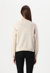 Suéter beige claro con mangas abullonadas texturizadas, escote y bajo acanalados, tejido de punto y corte relajado. Se lleva con pantalones negros.