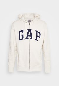 Felpa con cappuccio e zip di colore crema con il logo "GAP" in blu navy. Presenta due tasche frontali, polsini a costine e una texture morbida.