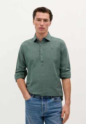 Camisetă verde, cu guler ascuțit, butonată, cu mânecile suflecate și un mic logo pe piept. Materialul pare moale și ușor texturat.