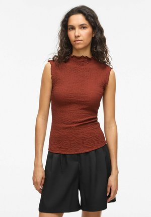 Femme aux cheveux ondulés foncés, portant un haut sans manches texturé couleur rouille et un short noir ample, debout devant un fond blanc uni.