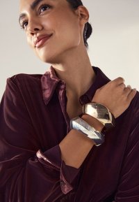 Gold- und Silberarmbänder mit geometrischem Design, übereinander getragen an einem Handgelenk, kombiniert mit einem burgunderfarbenen Samthemd mit Kragen- und Knopfdetaillierung.