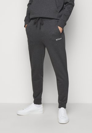 Person iført mørkegrå joggingbukser med en lille hvid logo på venstre lår og hvide sneakers, hænderne i lommerne, stående mod en ensfarvet baggrund.