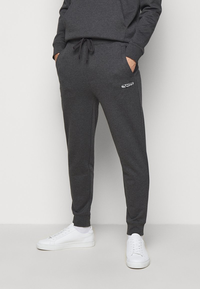 Personne portant un jogging gris foncé avec un petit logo blanc sur la cuisse gauche et des baskets blanches, les mains dans les poches, debout devant un fond uni.