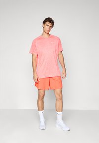 Joven de pie con camiseta atlética color coral y pantalones cortos, lleva puestos calcetas y zapatillas Nike blancas, con un fondo claro y simple.