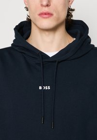 Tamsiai mėlynas džemperis su gobtuvu, pagamintas iš medvilnės, su raišteliu gobtuvui. Papuoštas baltu "BOSS" logotipu krūtinėje ir tekstūruota medžiaga.