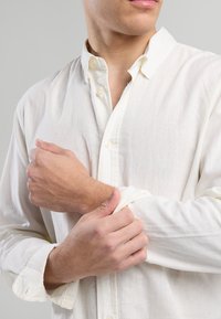 Uomo che aggiusta il polsino di una camicia bianca a maniche lunghe, inquadrato dalle spalle fino a metà torso, su uno sfondo grigio uniforme.