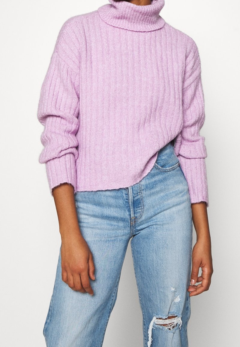 Personne portant un pull à col roulé côtelé lavande avec manches retroussées et un jean bleu clair déchiré, debout devant un fond uni.