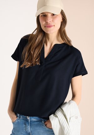 Frau trägt eine beige Kappe, ein schwarzes Kurzarm-V-Neck-Oberteil und blaue Jeans, hält eine weiße Nadelstreifenjacke über dem Arm.