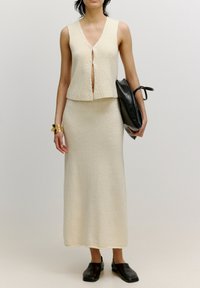 Cremfarbener Zweiteiler aus strukturiertem Stoff; ärmelloses Oberteil mit Frontöffnung und einem langen Rock. Schwarze Clutch-Tasche und Schuhe.