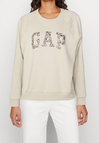 Felpa beige a girocollo con maniche raglan, caratterizzata da un logo "GAP" scintillante realizzato con dettagli metallici sulla parte anteriore.