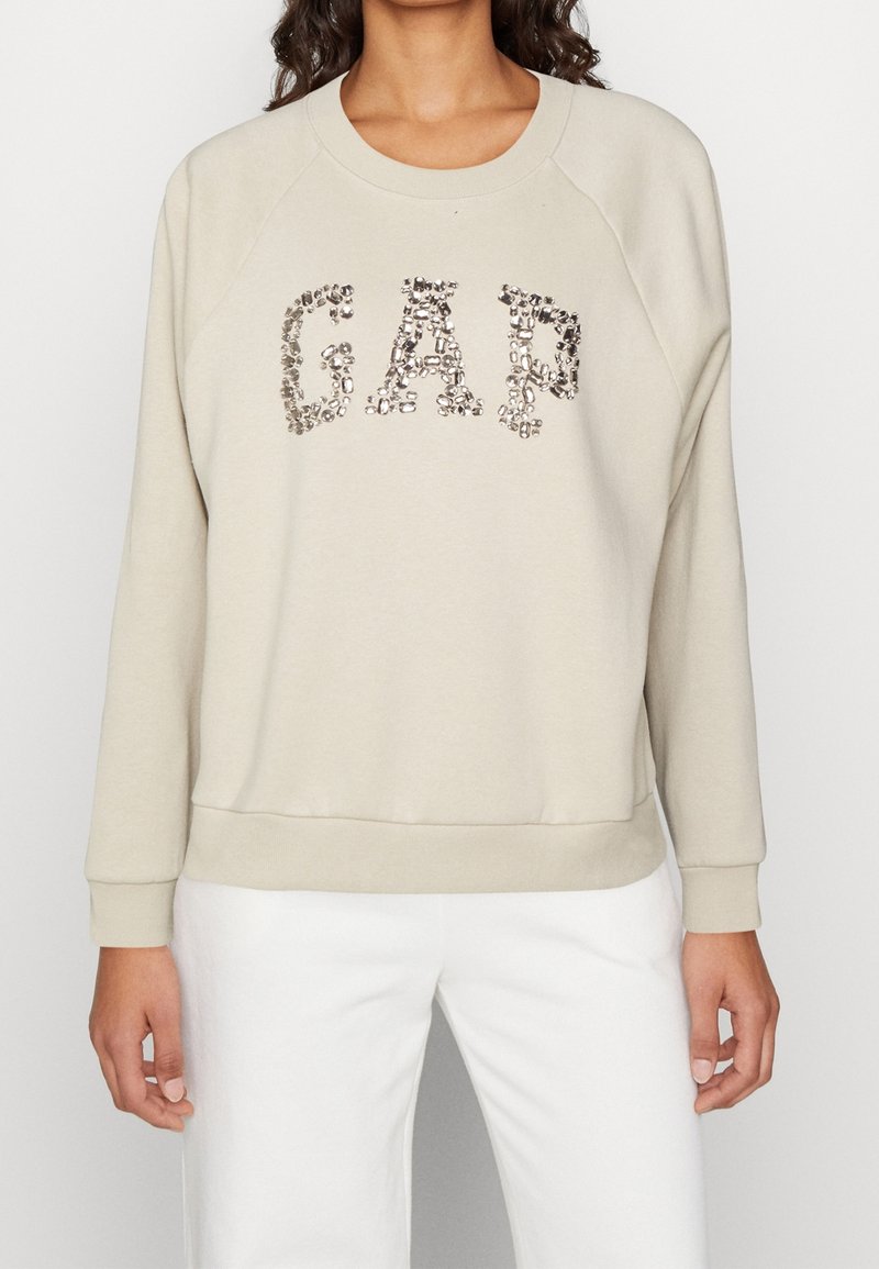 Felpa beige a girocollo con maniche raglan, caratterizzata da un logo "GAP" scintillante realizzato con dettagli metallici sulla parte anteriore.
