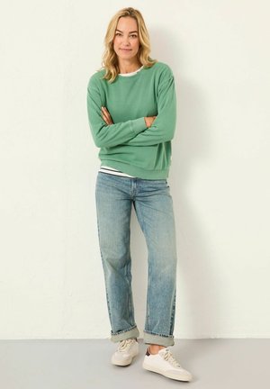 Woman with blonde hair wearing green sweatshirt, light blue rolled-up jeans, and white sneakers, standing against a white textured wall.