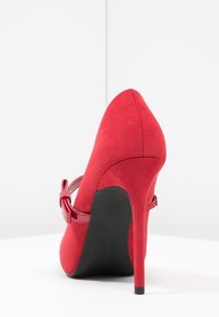 Red suede high heel shoe with a pointed toe and adjustable ankle strap, featuring a glossy red buckle accent. Black sole provides contrast.