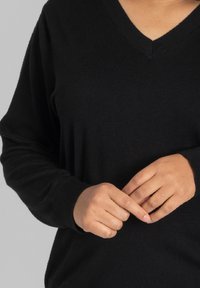 Schwarzer V-Ausschnitt-Pullover aus weichem Strickstoff mit lockerer Passform. Lange Ärmel und dezente gerippte Bündchen. Hände vorne positioniert.