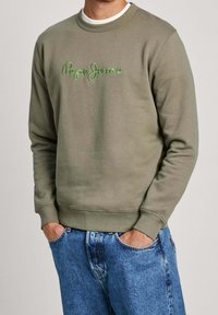 Mężczyzna w oliwkowej bluzie Pepe Jeans i niebieskich dżinsach z rękami w kieszeniach na jednolitym tle.