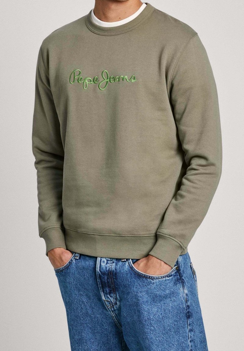Mężczyzna w oliwkowej bluzie Pepe Jeans i niebieskich dżinsach z rękami w kieszeniach na jednolitym tle.