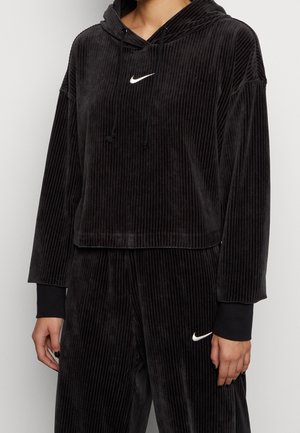 Osoba ubrana w czarną prążkowaną welurową bluzę z kapturem i pasujące spodnie z białymi logo Nike Swoosh na piersi i udzie.