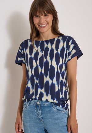 Femme souriante portant un top à manches courtes à motifs bleus et blancs et un jean en denim bleu clair sur un fond uni.