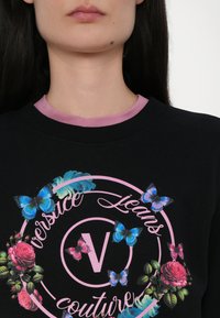 Čierna mikina s okrúhlym ružovým logom s písmenom "V" a kvetinovými grafikami, doplnená modrými motýľmi a vzormi ruží.