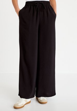 Osoba nosząca luźne, czarne spodnie o szerokich nogawkach z elastycznym pasem i sznurkiem, w połączeniu z białymi i beżowymi sneakersami na jednolitym tle.