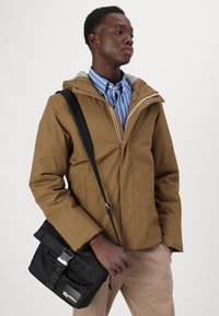 Giacca marrone con cappuccio, dal tessuto liscio, con chiusura a zip e fodera grigia, abbinata a una camicia a righe blu e a una borsa a tracolla nera.