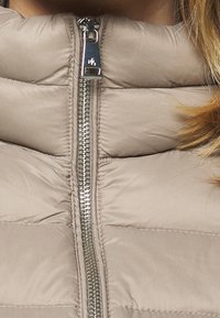 Beige vadderad jacka med quiltad struktur, utrustad med en silverfärgad dragkedja och ståkrage. Tyget ser slätt och lätt ut.