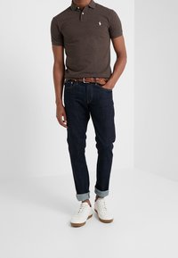 Man som bär en mörkbrun polo-shirt, mörkblå uppvikta jeans med flätat brunt bälte och vita sneakers, stående mot en vit blank vägg.