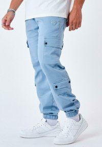 Pantalones cargo de color azul claro, hechos de una tela suave, con múltiples bolsillos, puños elásticos y un sutil detalle de logo. Combinados con zapatillas blancas.