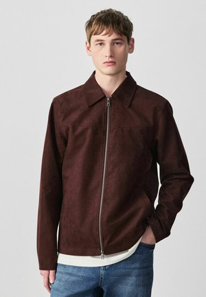 Junger Mann trägt eine dunkelbraune Jacke mit Reißverschluss und Kragen, darunter ein weißes Hemd und eine blaue Jeans, steht vor einem schlichten Hintergrund.