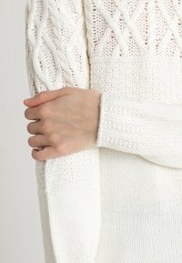 Main reposant sur l'avant-bras d'un pull blanc en tricot avec motif torsadé, montrant les textures détaillées du tricot et les poignets des manches.