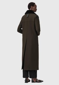 Casaco longo verde-oliva com gola de pele preta, rachas laterais e detalhes em pele nos ombros. Ajuste ajustado com um design elegante.