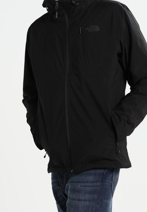 Mann trägt eine schwarze North Face Jacke mit Reißverschluss, die Hände in den Taschen und blaue Jeans vor einem einfarbigen Hintergrund.