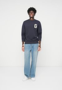 Marineblå genser med "G"-logo, sammen med lyseblå vide jeans og hvite joggesko. Glatt stoff og komfortabel passform.