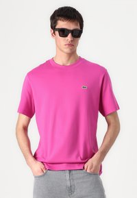Přiléhavé růžové bavlněné tričko s krátkým rukávem, kulatým výstřihem a malým zeleným logem Lacoste na hrudi. Nosené s šedými džíny.