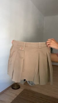 Bēša plisēta svārku kleita, izgatavota no auduma ar gludu tekstūru, ar birku, jostu cilpām un vidēja izmēra jostasvietu.