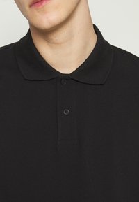 Polo noir avec un col classique, une patte à deux boutons et un tissu doux et texturé. Vue en gros plan montrant les détails du matériau et des coutures.