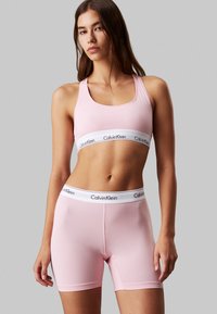 Calvin Klein ružičasti atletski bralet s bijelom logom trakom, uparen s ružičastim hlačama koje imaju odgovarajući logotip na pojasu. Glatka tkanina, prilagođeni dizajn.