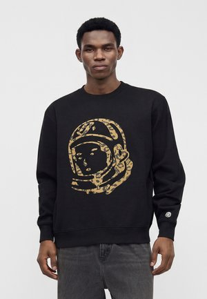 Jeune homme portant un sweat-shirt noir avec un grand motif de casque d'astronaute à imprimé léopard sur le devant et un jean foncé.