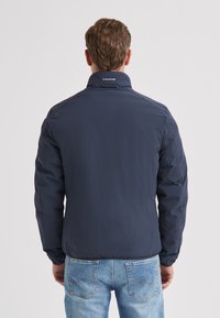 Marineblaue leichte Jacke mit hohem Kragen, glatter Textur und elastischen Bündchen. Verfügt über ein Markenlogo auf der Rückseite und ein minimalistisches Design.
