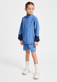 Ljusblå träningsjacka med marinblå ärmar, dragkedjestängning och elastiska muddar, matchad med shorts och vita sneakers.