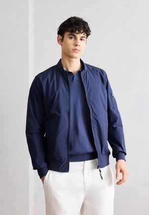 Mladý muž s kučeravými vlasmi, oblečený v námornícky modrej bunde na zips, námornícky modrom svetri a bielych nohaviciach, stojaci pred jednoduchým svetlým pozadím.