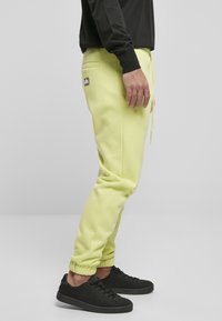 Pantaloni de trening galbeni deschis, cu talie elastică și manșete, având o buzunar lateral și un patch cu logo negru. Asortați cu adidași negri.